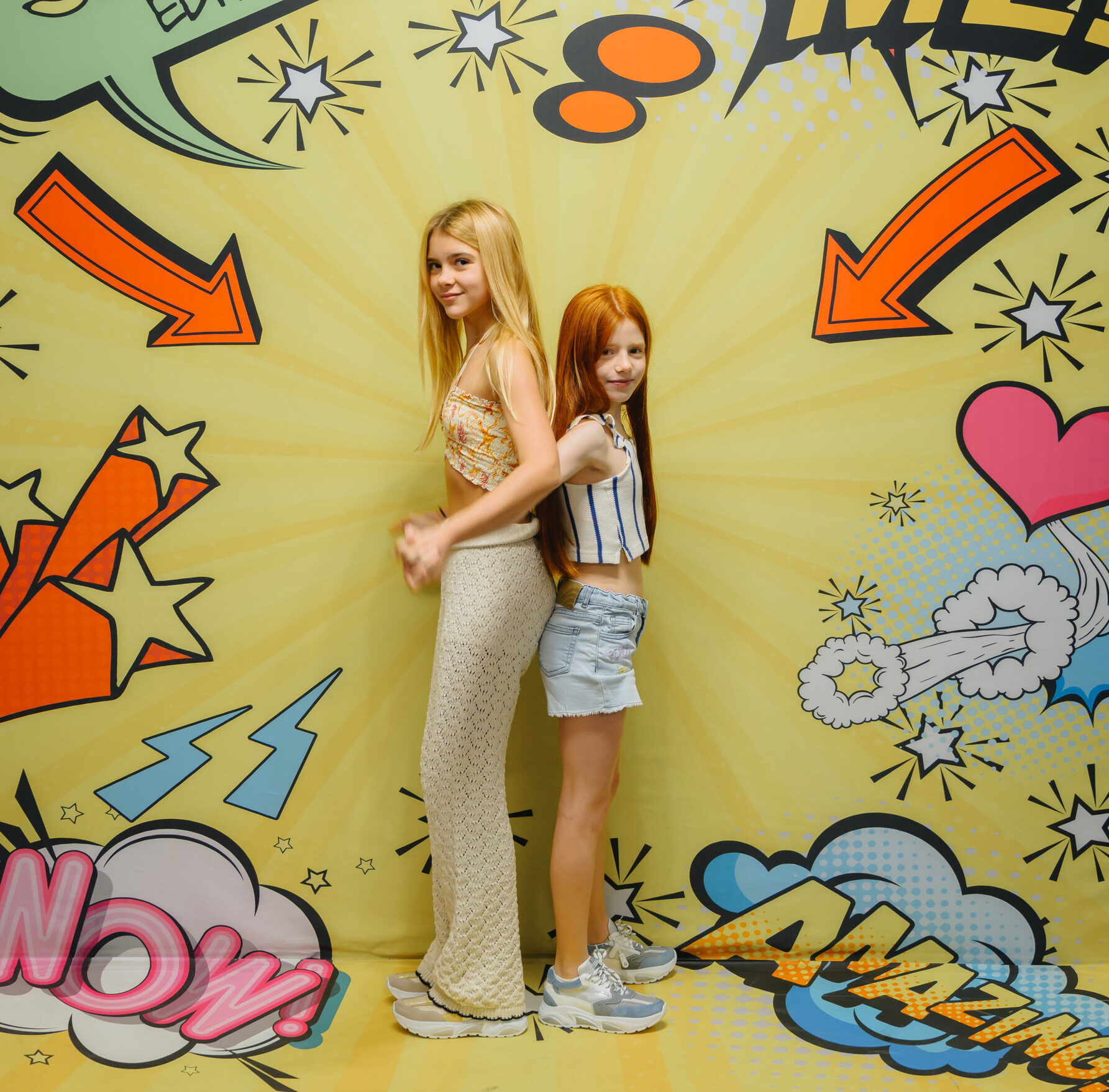 Two girls, a blonde and a redhead, posing back-to-back in GIGAshoes sneakers in front of a yellow comic book style background with speech bubbles like "SPECIAL EDITION" and "FOLLOW ME!