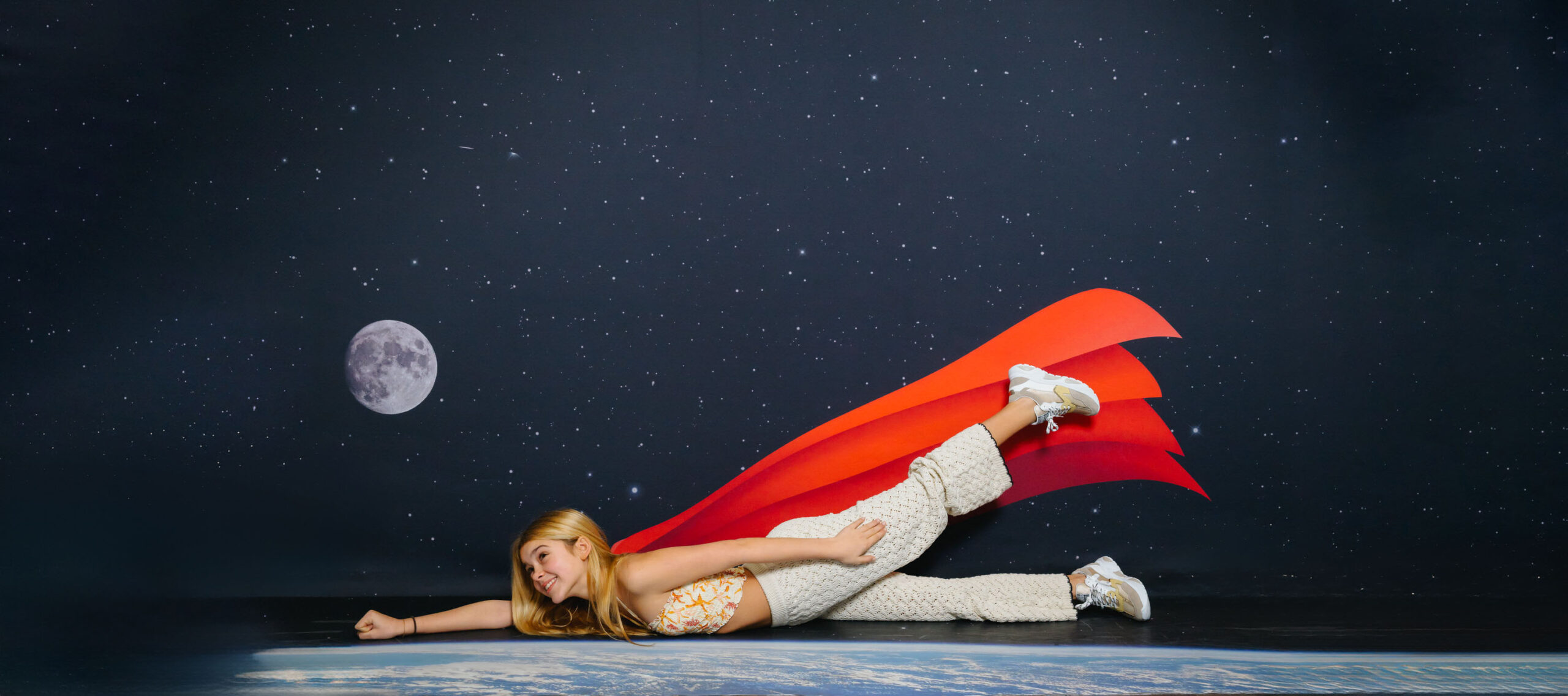 Teenager in GIGAshoes sneakers flying over Earth in a starry space setting with a red cape.