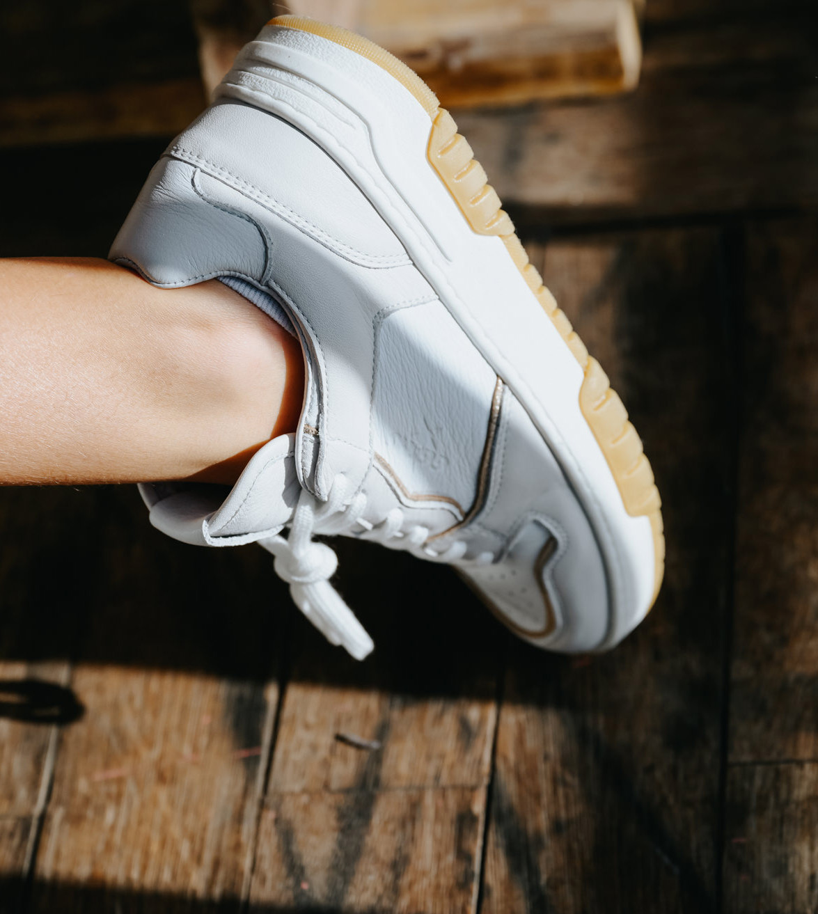 Close-up of a white leather GIGAshoes sneaker with a tan gum sole, worn by a child sitting on a wooden bench in natural sunlight.