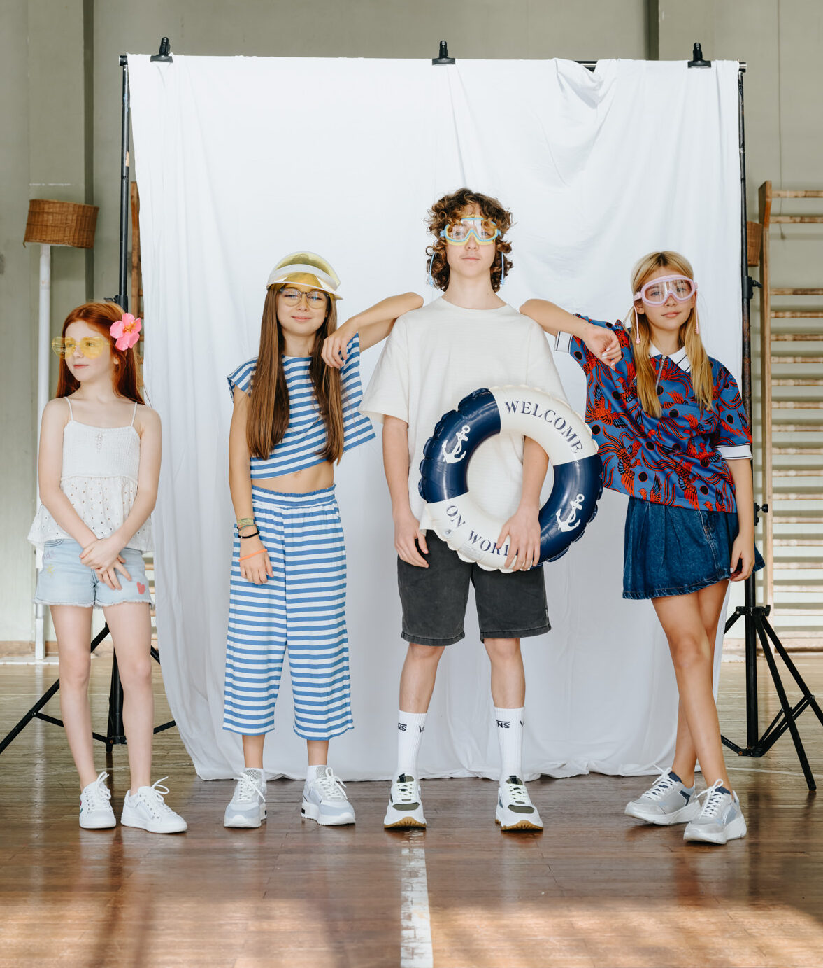 Group of four kids and teenagers wearing GIGAshoes white and grey sneakers, holding a lifebuoy and wearing summer goggles in an indoor gym setting.