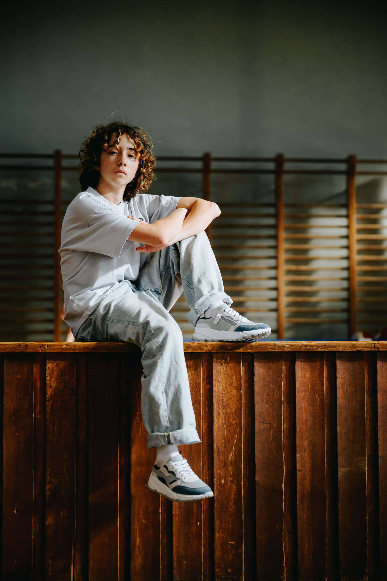 Teen boy with curly hair sitting on a wooden bench in an old-school gym, wearing light blue jeans and grey and white GIGAshoes sneakers with a thick sole.