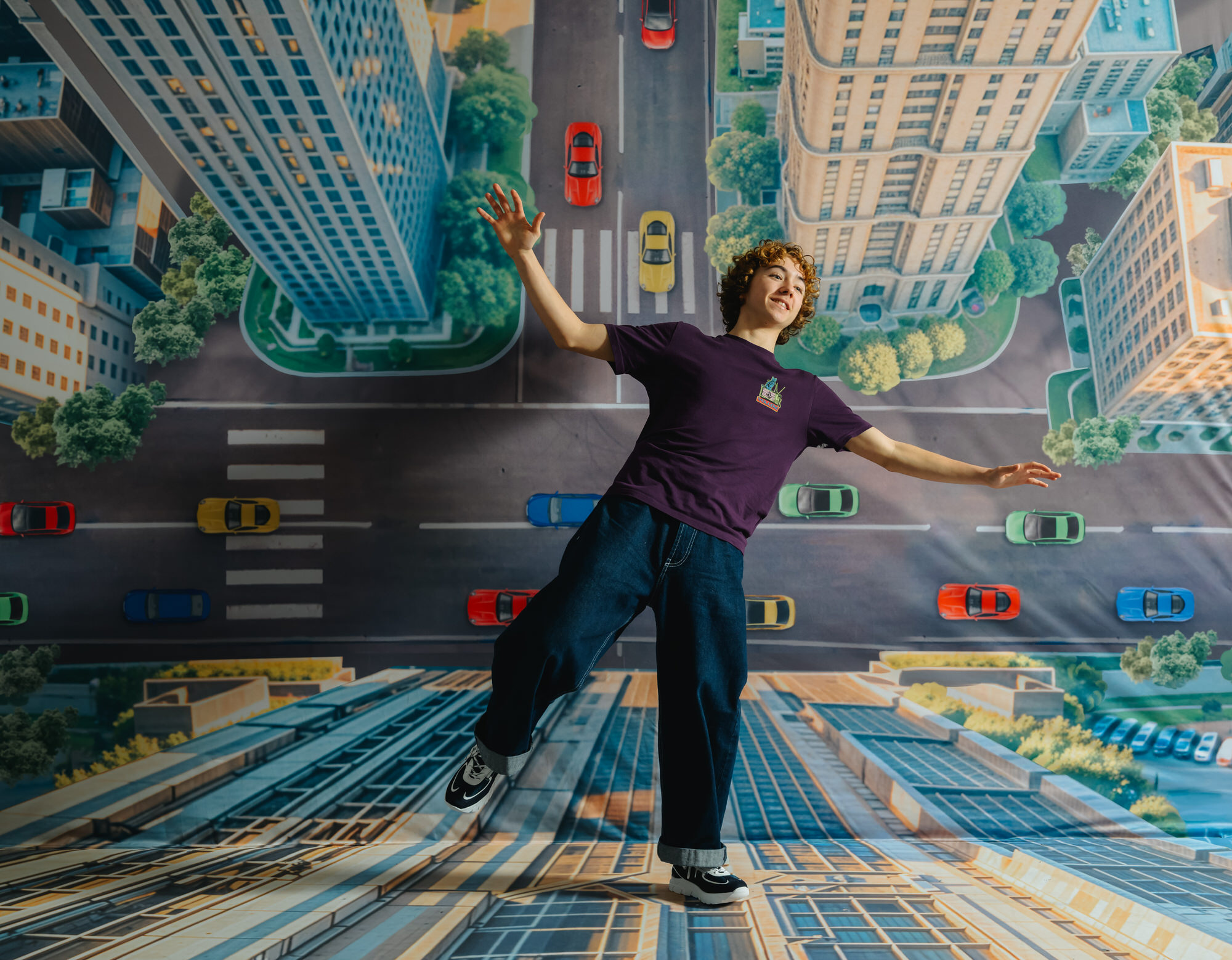 Teen boy with curly hair, purple t-shirt, baggy jeans, and GIGAshoes sneakers, appearing to walk down the side of a building on a painted city street with cars below.