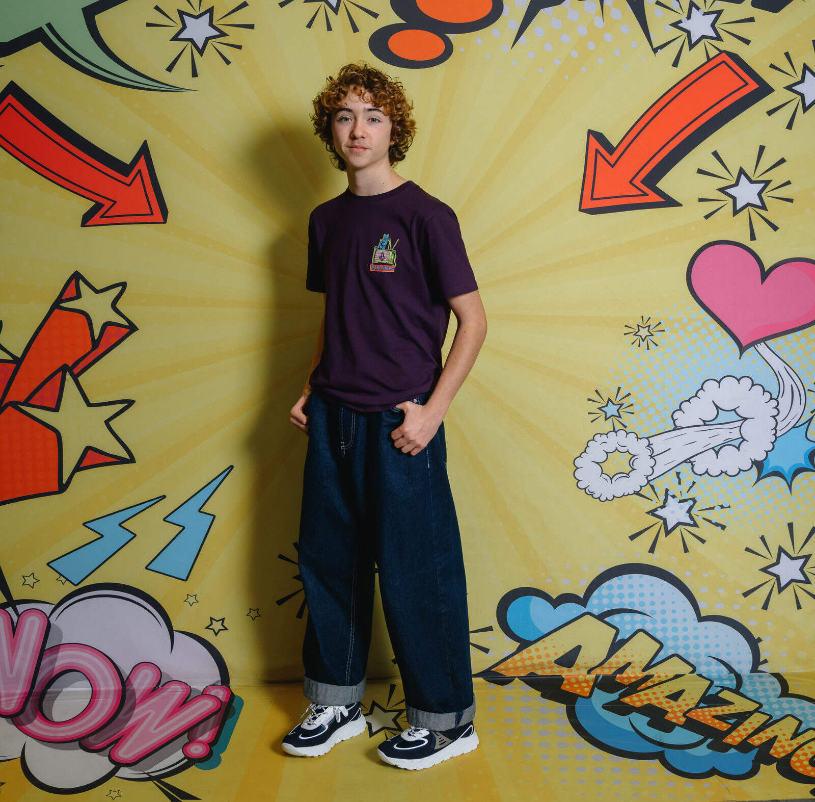Teenager posing in front of a yellow comic-style background with speech bubbles, wearing GIGAshoes grey sneakers and baggy jeans.